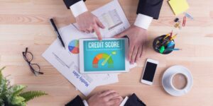 Image of two persons hands on a table with one showing the credit score displayed on a tab.Few documents a mobile and stationery items are kept around it.