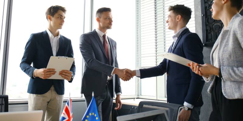 A group of businessmen standing in a conference room with two of them shaking hands after a strategic tieup.