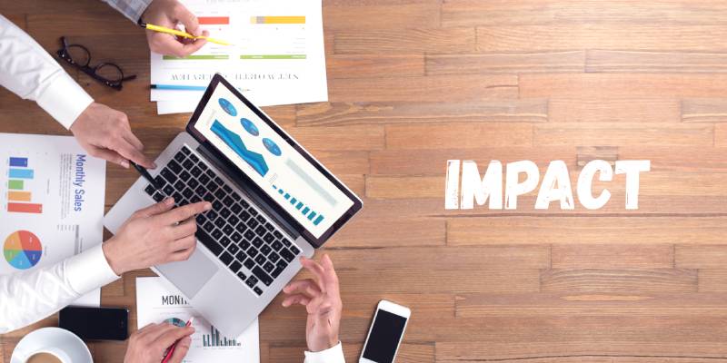 A group of persons discussing financial matters using graphs and data on printouts and laptop where the word impact is mentioned on the same table near it.