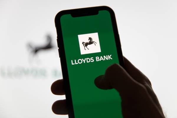 A persons hand holding a smartphone displaying the logo of LloydsBank on a green background and a blurred logo of the same behind the image.