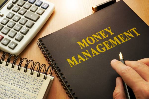 A person pointing his pen towards the term money management written on a book kept near a calculator and a notepad with some transaction details mentioned on it.