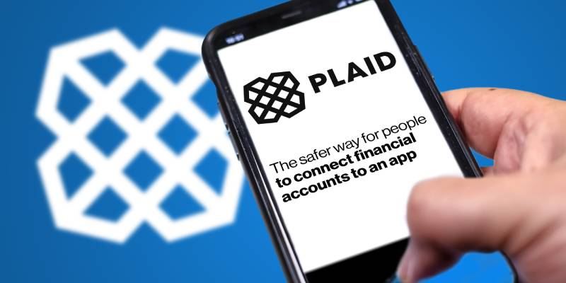 A hand holding a phone with Plaid homepage on screen with plaid logo blurred on a blue background behind it.
