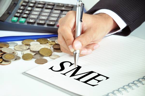 A hand with pen pointing to the word SME written on a paper with a few coins and a calculator beside it.
